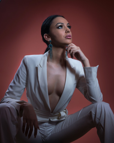 年轻美丽的黑发女人穿着雅致的白色西装，裤子坐着。迷人的黑头发女孩摆姿势，演播室拍摄。穿着白色西服的优雅女士，外套，领口下垂，怀旧