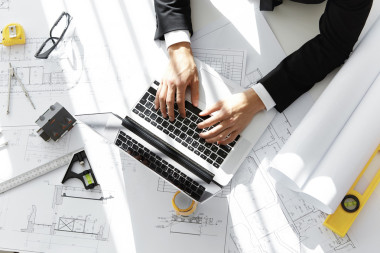 建筑師或承包商坐在辦公桌前與筆記本電腦，草圖，規(guī)模模型房子，藍圖卷和尺子，輸入數(shù)據(jù)，而在他的辦公室工作新的住房項目