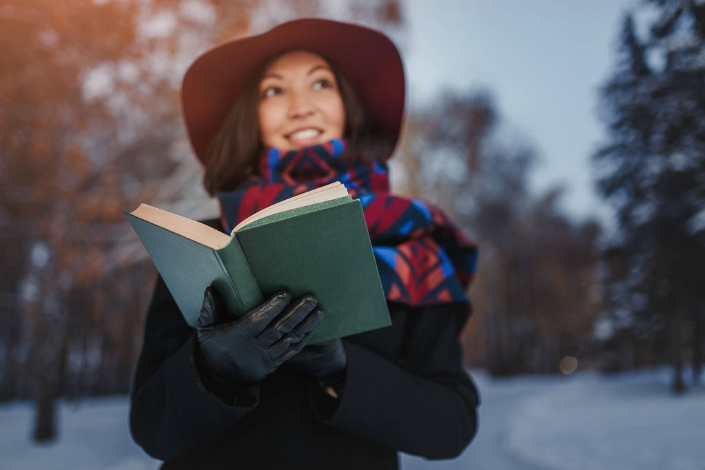 红色帽子持有和阅读诗歌书在冬季森林背景的年轻女子 红色帽子持有和阅读诗歌书在冬季森林背景的年轻女子