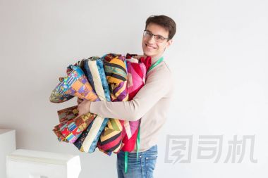 英俊的年輕男裁縫在白色背景上拿著一疊折疊的顏色拼布絎縫被。許多彩色毛毯樣式拼湊而成的男人微笑著，手工手中