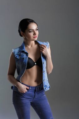在灰色的背景上的牛仔马甲性感年轻的黑发女人的画像