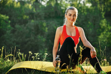 在紅色的頂部和黑色褲子的年輕金發(fā)女郎坐在那兒，一瓶水在大自然中健身的黃色地毯上
