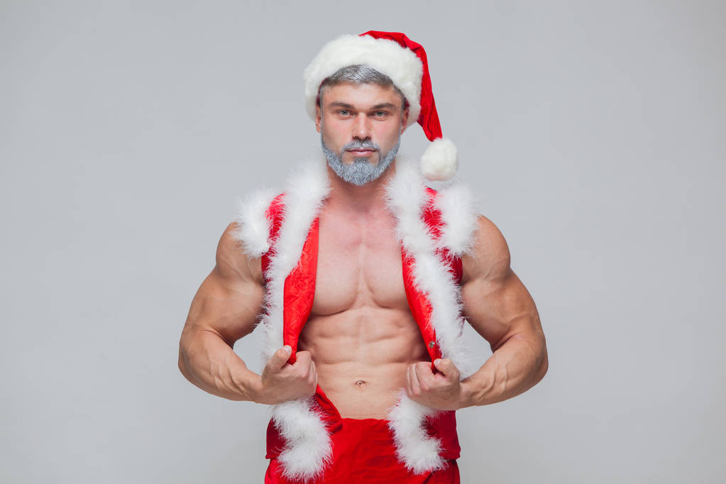圣诞节。性感的圣诞老人。年轻的肌肉男穿圣诞老人帽子展示他的肌肉 圣诞节。性感的圣诞老人。年轻的肌肉男穿圣诞老人帽子展示他的肌肉