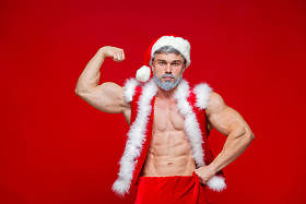 圣诞节。体育，活动。性感的圣诞老人。年轻的肌肉男穿圣诞老人帽子展示他的肌肉。红色背景上的孤立。演示大二头