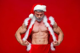 圣诞节。体育，活动。性感的圣诞老人。年轻的肌肉男穿圣诞老人帽子展示他的肌肉。红色背景上孤立