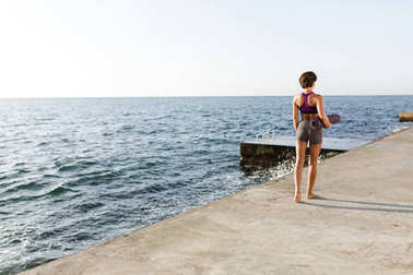 年輕女子在海邊散步時手持瑜伽墊, 手里拿著深色短發(fā)。美麗的女士在運動的頂部和短褲站在她的背部與海景色的背景