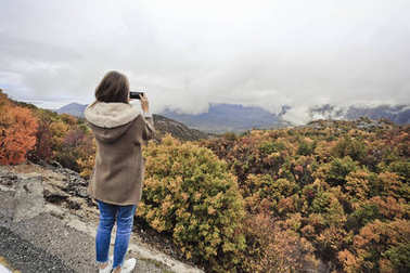 Trandy 的女孩站在后面, 拍著在手機(jī)上多云的天空下的陰雨的秋天山的風(fēng)景。旅游理念