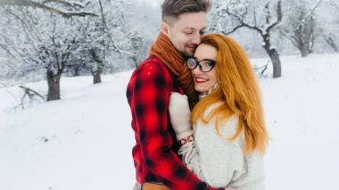 在白雪皑皑的森林里拥抱恋人的可爱肖像。带着眼镜的美丽微笑的红女人