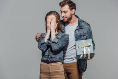英俊微笑的年轻人拿着礼品盒, 而他的女朋友闭上眼睛孤立灰色