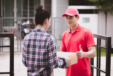 年輕的后勤職業(yè)概念。快樂送貨的人把他的包給客戶在家里。在真正的房子。二十歲代初, 亞洲華人身穿紅色馬球衫和紅帽牛仔褲