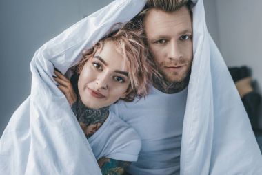 年轻的纹身情侣在卧室的白色毯子下看着相机