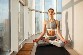 在运动的顶部和绑腿坐在莲花姿势的周到的女士肖像和冥想的瑜伽垫在家里与大美丽的窗口背景