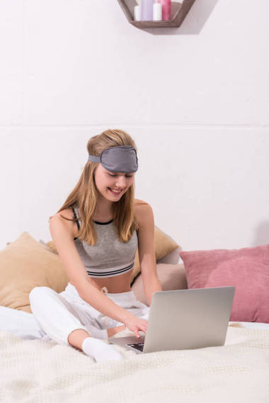 美麗的年輕女子與睡眠面具的頭部工作與筆記本電腦, 而坐在床上的早晨