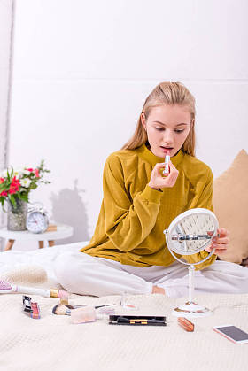 年轻妇女在床上涂抹口红在家里, 看着镜子