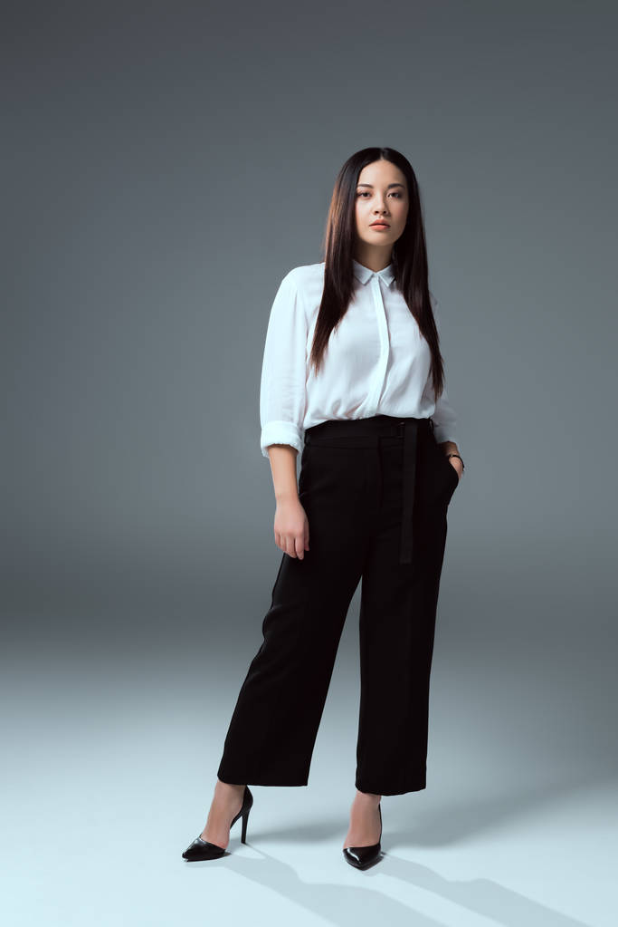 美丽的亚洲女孩的全长视图站在口袋里, 看着在灰色的摄像头