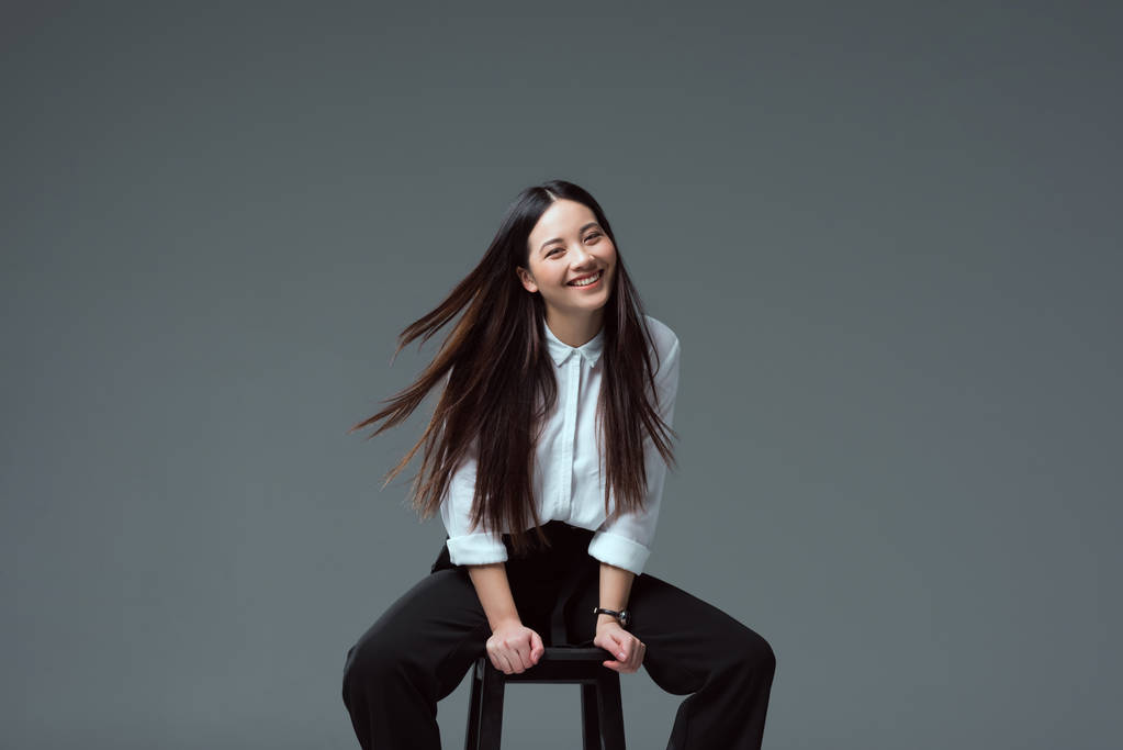 迷人的亚洲女孩坐在椅子上, 微笑着在镜头上的灰色隔离