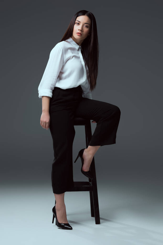 美丽时尚的亚洲女孩的全长视图摆在椅子上, 看着在灰色的摄像头