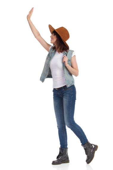 身穿褐色帽子、牛仔褲背心和靴子的年輕女子正站著揮舞著手。側(cè)面視圖