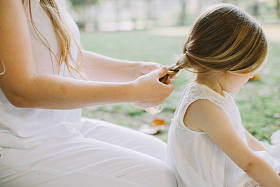 美丽的年轻母亲的肖像, 使她可爱的小女儿在公园编织辫子