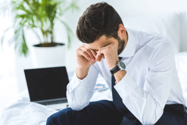 疲憊的商人坐在床上與筆記本電腦