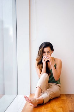 年輕的亞洲華人婦女在早晨喝一杯透明的不倒翁的健康混合奶昔。她坐在她公寓的窗戶旁, 陽(yáng)光灑在屋里。她穿著舒