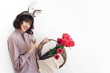 复活节狩猎概念。美丽的时尚女孩抱着篮子与郁金香和复活节彩蛋站在白色背景