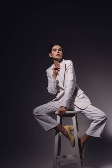 时尚的年轻女子穿着白色西装坐在椅子上的黑暗背景