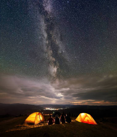 四名徒步旅行者坐在兩個橙色帳篷營地, 晚上享受美麗的星空和銀河。在遠(yuǎn)處的城市的燈光坐落在山之間