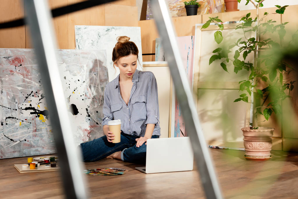 有选择的焦点, 美丽的年轻艺术家拿着咖啡去和使用笔记本电脑在工作室 有选择的焦点, 美丽的年轻艺术家拿着咖啡去和使用笔记本电脑在工作室