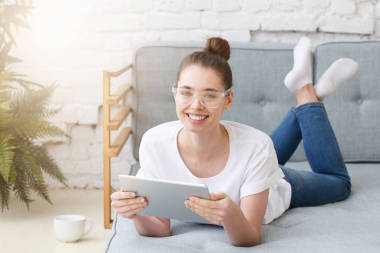 快樂美麗的年輕女子戴著透明的眼鏡, 躺在沙發(fā)上, 在家里用數碼片, 笑得很廣