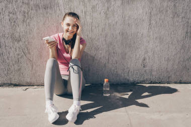 快樂的女孩坐在戶外, 鍛煉后休息, 并獲得力量, 持有智能手機