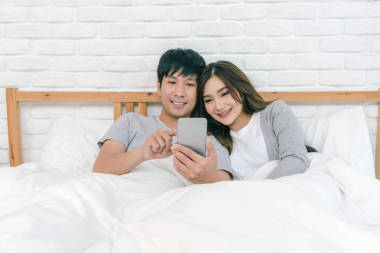 幸福的亞洲夫婦使用技術(shù)智能手機(jī)在床上的臥室在家里, 情侶和生活方式的概念