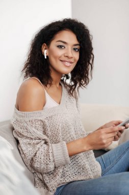 戴著耳機(jī)的黑卷發(fā)的年輕女士的肖像坐在沙發(fā)上, 手里拿著平板電腦, 高高興興地看著家里的相機(jī)