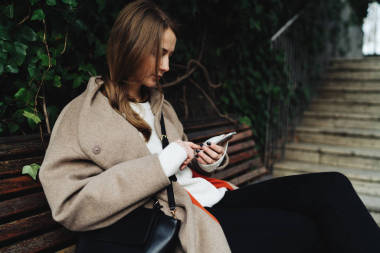 年輕女子穿著時髦的服裝, 而坐在戶外木凳上使用智能手機