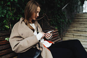 年轻女子穿着时髦的服装, 而坐在户外木凳上使用智能手机