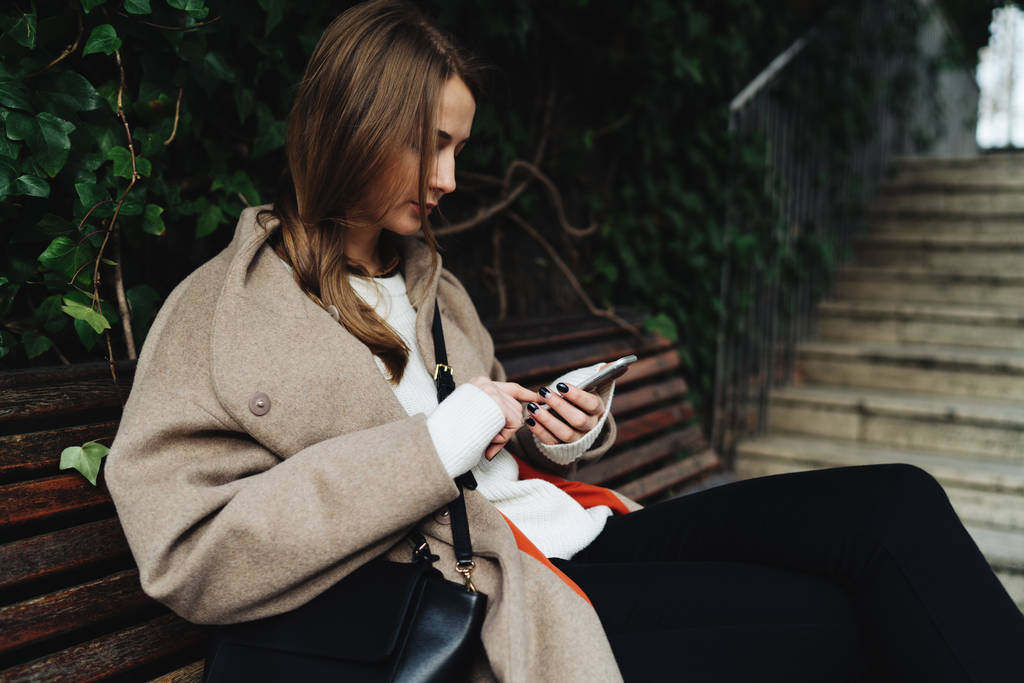 年轻女子穿着时髦的服装, 而坐在户外木凳上使用智能手机 年轻女子穿着时髦的服装, 而坐在户外木凳上使用智能手机