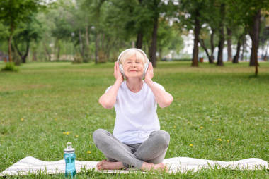 快樂的老太太聽音樂與耳機坐在瑜伽墊與瓶水在公園里 