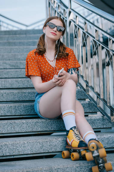 有魅力的女孩在太阳镜和轮滑鞋坐在楼梯上, 听音乐与智能手机