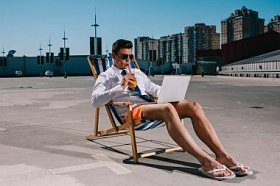 英俊的年轻商人在短裤工作与笔记本电脑, 而坐在太阳躺椅在停车和喝鸡尾酒