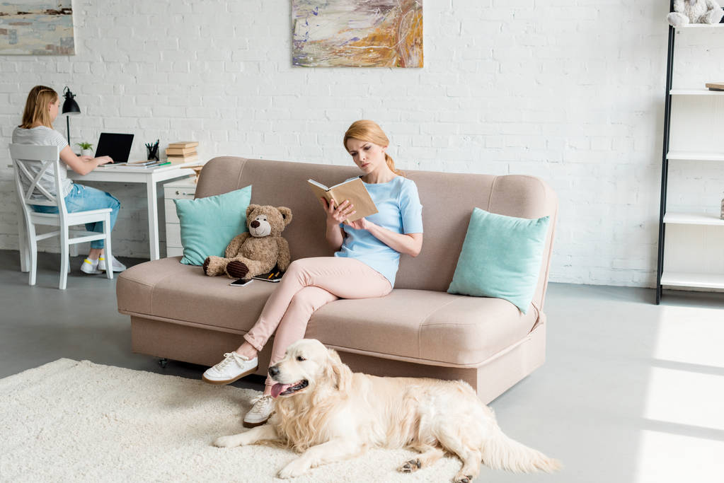 集中的母亲看书和坐在沙发上, 而她的女儿使用笔记本电脑 集中的母亲看书和坐在沙发上, 而她的女儿使用笔记本电脑