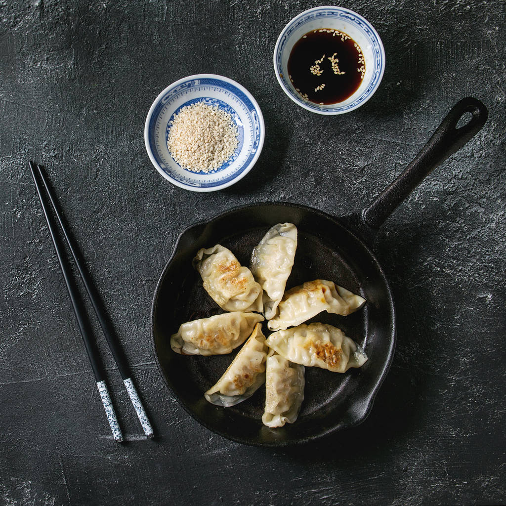 亚洲饺子 Gyozas 锅贴炒在铸铁锅上, 配有筷子和一碗酱油, 上面有黑色的纹理背景。顶部视图, 空间