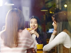 三快乐美丽的年轻亚洲妇女坐在桌边聊天在咖啡馆或茶馆, 通过窗口玻璃拍摄
