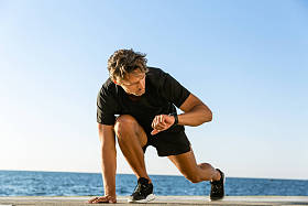 英俊的成人短跑运动员看健身跟踪, 而站在开始的位置运行在海边