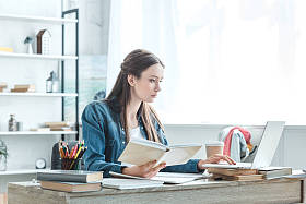 重点青少年女孩拿着书和使用笔记本电脑, 而在家学习