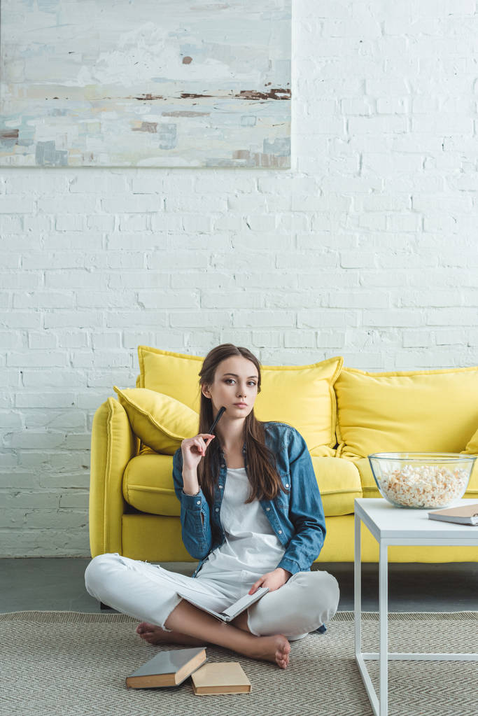 有吸引力的女孩拿着书, 看着相机, 而坐在地毯上, 在家学习 有吸引力的女孩拿着书, 看着相机, 而坐在地毯上, 在家学习