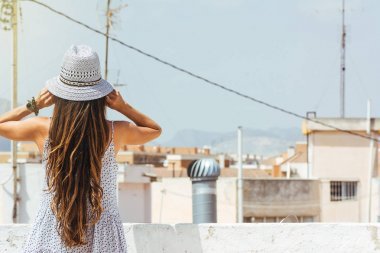 長頭發(fā)的女人戴著帽子看著地平線上的城市, 從后面, 穿著在一個夏天的禮服
