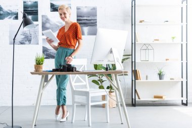 時尚女性攝影師使用數(shù)字平板電腦和照片相機在家里辦公室附近的表