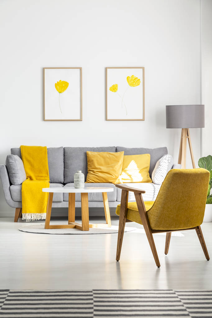 海报与黄色的花朵挂在明亮的客厅内的灰色沙发上与复古扶手椅和木制咖啡桌。真实照片 海报与黄色的花朵挂在明亮的客厅内的灰色沙发上与复古扶手椅和木制咖啡桌。真实照片