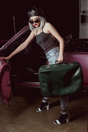 时尚的金发女郎在贝雷帽和太阳镜打开老式汽车和持有老式手提箱