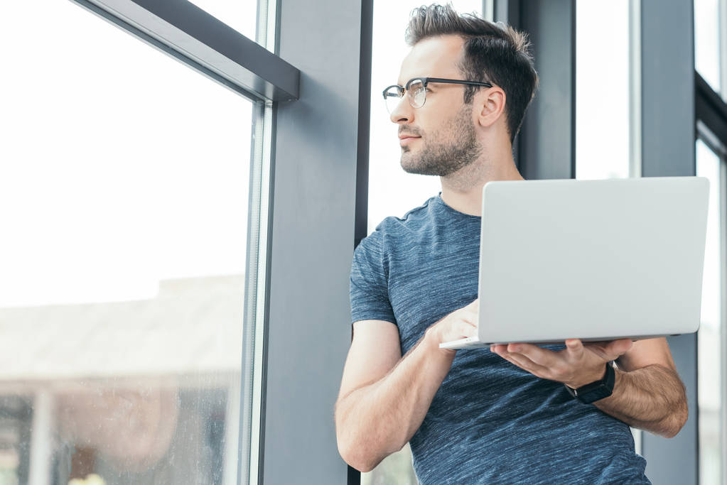 英俊的年轻男子在眼镜持有笔记本电脑和看窗口 英俊的年轻男子在眼镜持有笔记本电脑和看窗口